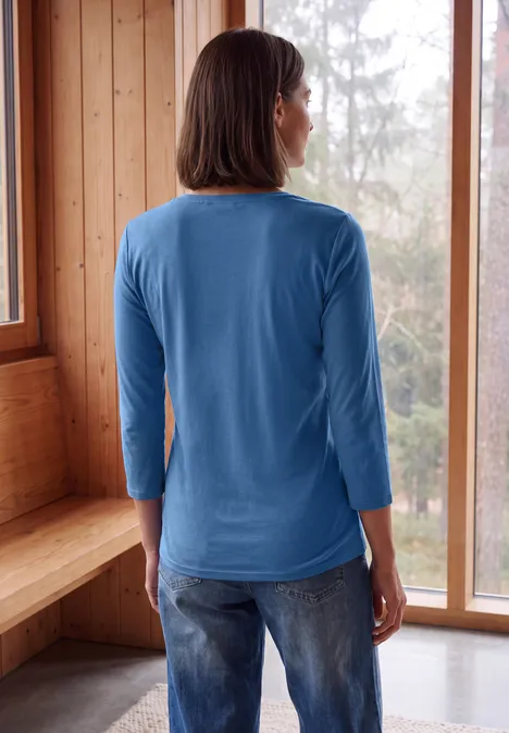 3/4-Arm Shirt in Unifarbe mit V-Neck north blue Frau von hinten in blauem Shirt und Jeans, steht in hellem Holzraum vor großen Fenstern mit Blick auf Wald.