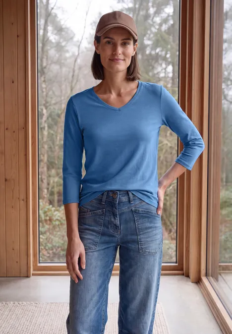 3/4-Arm Shirt in Unifarbe mit V-Neck north blue Frau in blauer V-Neck-Strickbluse, Jeans und Cap steht lächelnd vor raumhohen Fenstern aus Holz.