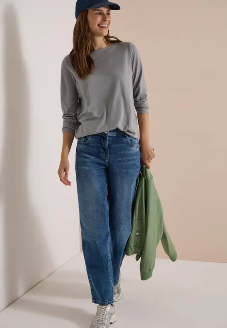 Langarm Shirt mit U-Boot-Ausschnitt concrete grey melange Eine Frau in grauem Shirt, blauen Jeans und Baseballcap lehnt entspannt an einer Wand und hält eine grüne Jacke.