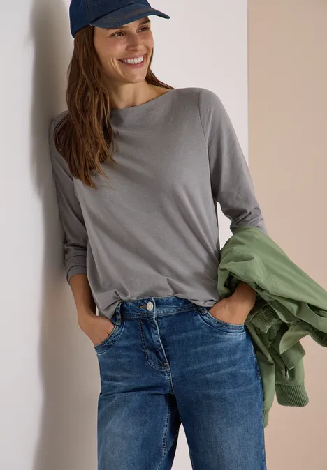 Langarm Shirt mit U-Boot-Ausschnitt concrete grey melange Frau lehnt lächelnd an Wand, trägt Jeans, graues Shirt, blaue Cap und hält grüne Jacke.