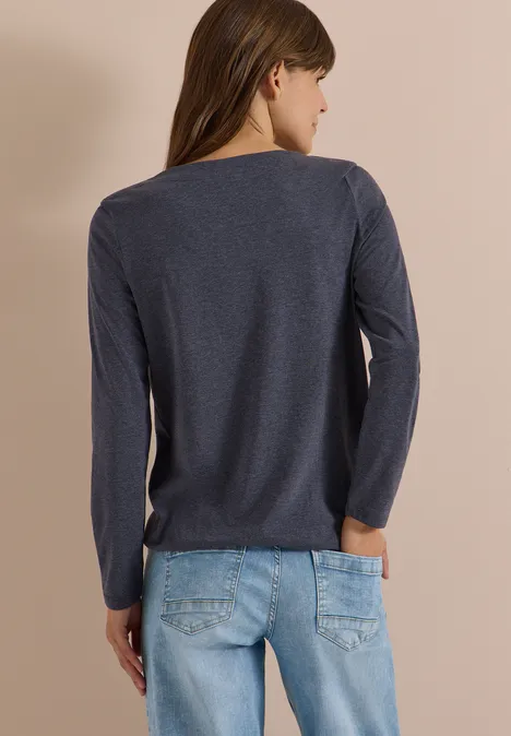 Vrouw van achteren in donkerblauw lang shirt en lichtblauwe spijkerbroek voor een beige achtergrond.