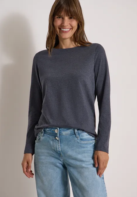 Vrouw in grijs shirt met lange mouwen en lichtblauwe jeans lacht voor een lichte achtergrond.