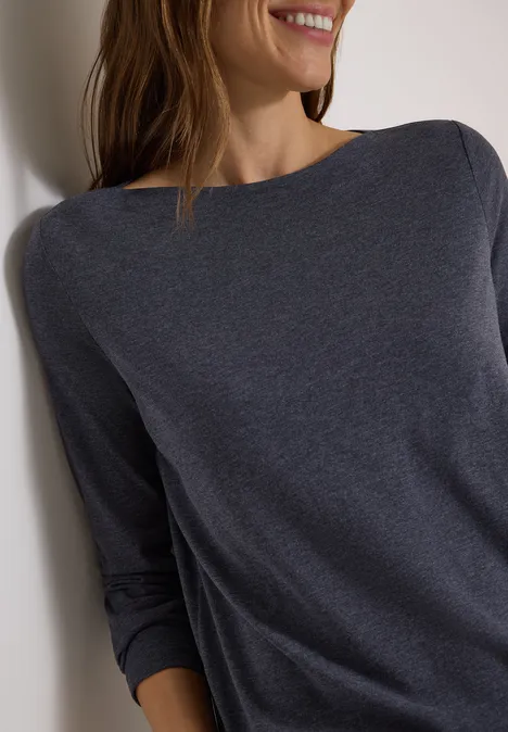 Vrouw leunt tegen witte muur, draagt donkergrijs shirt met lange mouwen en glimlacht.