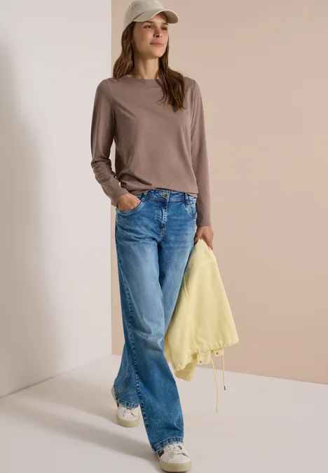 Jonge vrouw met pet, bruin shirt, wijde blauwe jeans, witte sneakers, houdt een gele jas vast.