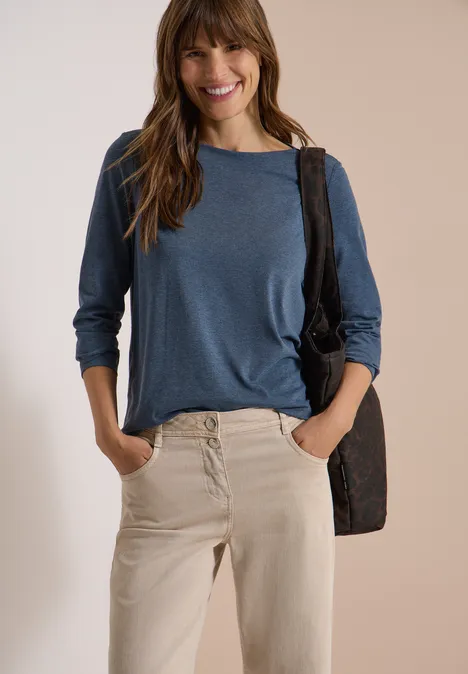 Langarm Shirt mit U-Boot-Ausschnitt moonlight blue melange Frau in beiger Hose und blauem Langarmshirt, lächelnd, Hände in den Taschen, mit schwarzer Umhängetasche.
