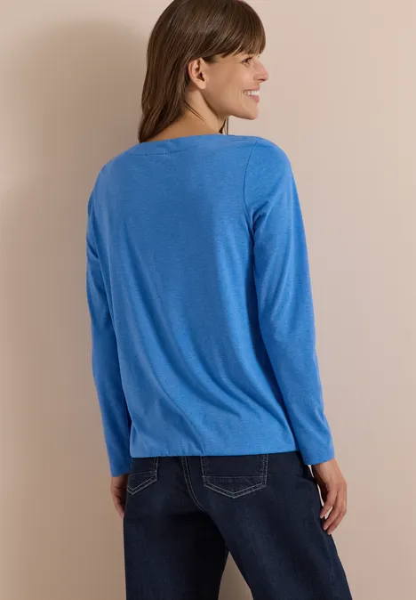 Femme de dos portant un top bleu à manches longues et un jean devant un mur clair.