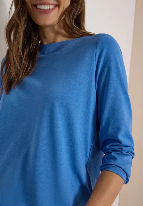 Une femme porte un haut bleu à manches longues, style décontracté, et sourit légèrement.