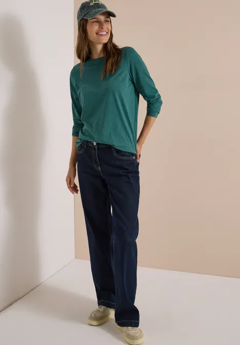 Langarm Shirt mit U-Boot-Ausschnitt dark fir green melange Frau mit grüner Langarmshirt, Jeans und Cap, lächelnd in neutralem Raum stehend.