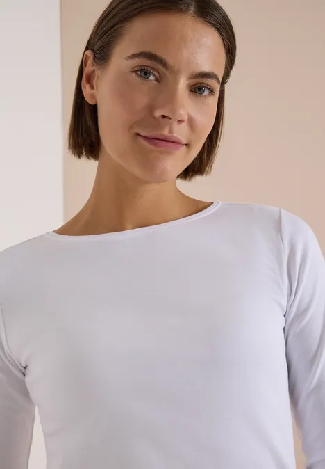 Langarm Shirt mit U-Boot-Ausschnitt White Junge Frau mit schulterlangem braunen Haar, weißem Shirt und leichtem Lächeln vor hellem Hintergrund.