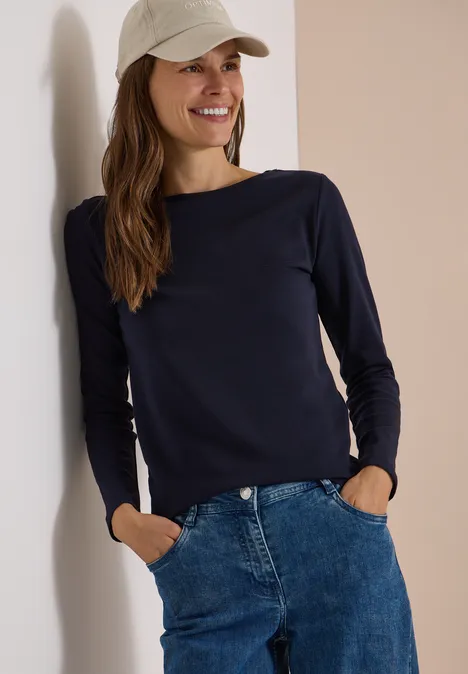 Langarm Shirt mit U-Boot-Ausschnitt urban dark blue Eine lächelnde Frau lehnt an einer Wand, trägt blaue Jeans, ein dunkelblaues Langarmshirt und eine beige Baseballkappe.