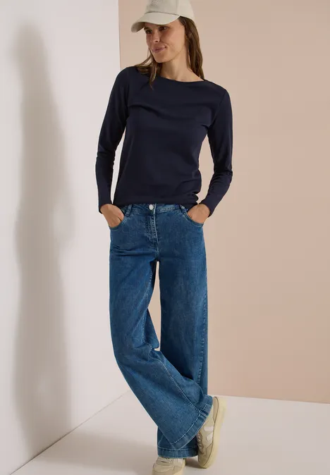 Langarm Shirt mit U-Boot-Ausschnitt urban dark blue Eine Frau mit weißer Cap, schwarzem Pullover, weiten Jeans und Sneakern lehnt an einer Wand.