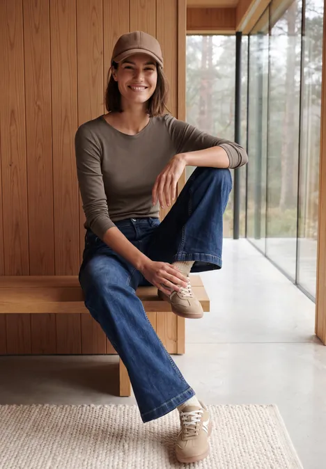 Langarm Shirt mit U-Boot-Ausschnitt urban taupe Frau in Jeans, Pullover und Cap sitzt lässig auf einer Holzbank vor großen Panoramafenstern.