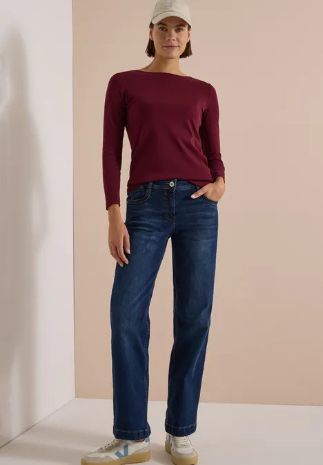 Langarm Shirt mit U-Boot-Ausschnitt red wine Frau in dunkelrotem Langarmshirt, blauen Jeans, weißen Sneakern und beige Cap vor neutralem Hintergrund.