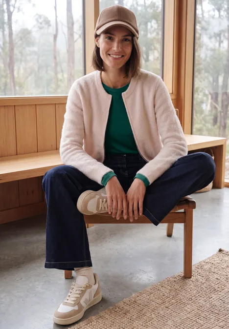 Langarm Shirt mit U-Boot-Ausschnitt dark fir green Frau sitzt lächelnd auf Holzbank, trägt Baseballcap, hellen Cardigan, grünes Top, Jeans und bequeme Sneaker.