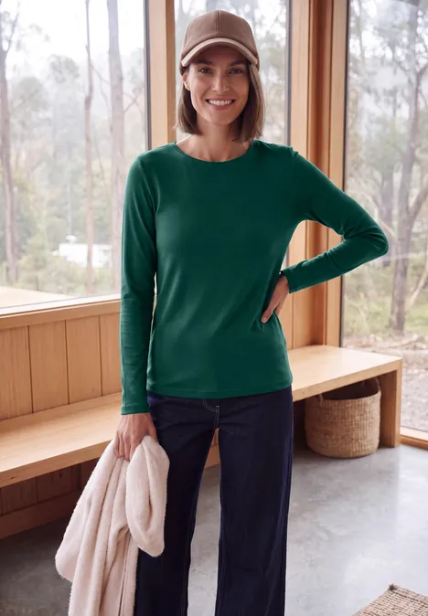 Langarm Shirt mit U-Boot-Ausschnitt dark fir green Lächelnde Frau in grünem Langarmshirt, Jeans und Baseballcap hält hellen Pullover vor Holzfenster.