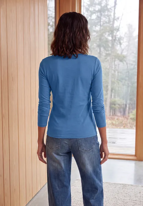 Langarm Shirt mit U-Boot-Ausschnitt north blue Person von hinten in blauem Oberteil und Jeans, stehend an Fenster in hellem Holzraum.