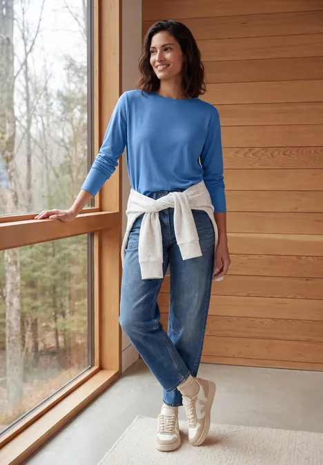 Langarm Shirt mit U-Boot-Ausschnitt north blue Frau in blauem Pullover und Jeans mit hellem Pullover um die Taille, lächelnd am Fenster
