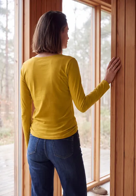 Langarm Shirt mit U-Boot-Ausschnitt golden honey Frau in gelbem Pullover und Jeans steht an einer Glas-Schiebetür und schaut nach draußen.