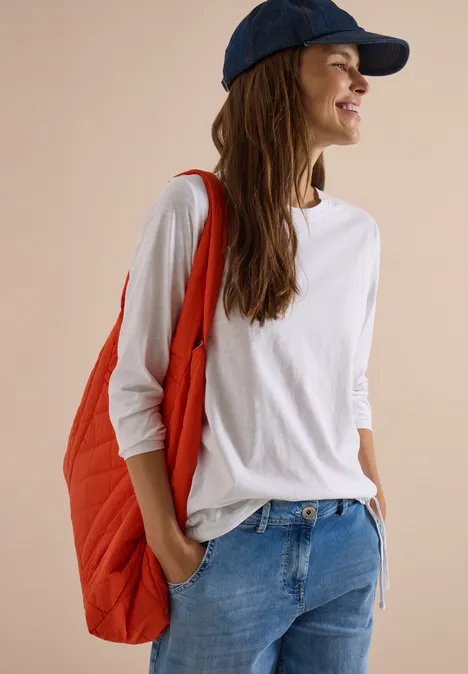 Femme souriante en t-shirt blanc, jean, sac rouge et casquette bleue, regardant sur le côté.