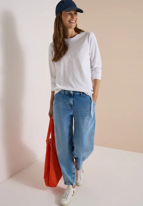Une jeune femme en jean ample, t-shirt blanc et casquette bleue porte un sac rouge et baskets blanches.