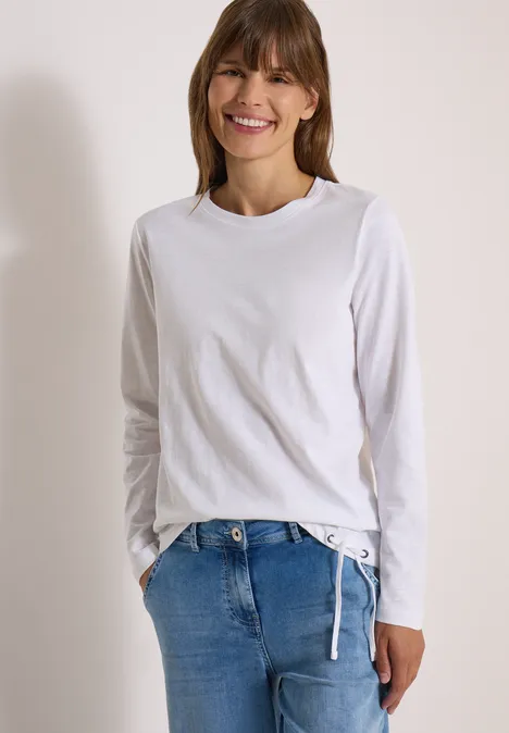 Une femme souriante en jean et t-shirt à manches longues blanches, posant devant un fond clair.