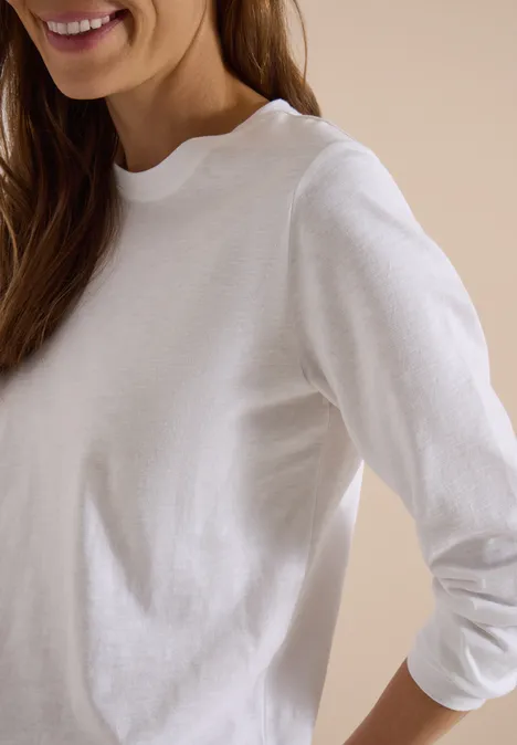 Femme portant un t-shirt à manches longues blanc, seul le buste et un sourire discret sont visibles.