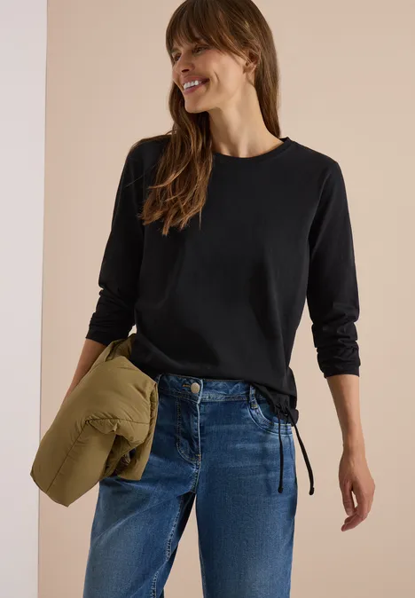 Femme en t-shirt noir à manches longues et jean, tenant une veste kaki pliée, souriante.