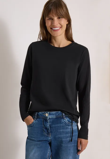 Femme souriante en pull noir à manches longues et jean bleu, mains dans les poches.