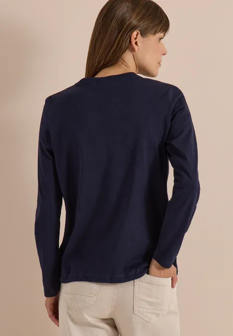 Femme de dos portant un haut bleu marine à manches longues et un pantalon clair, fond neutre.