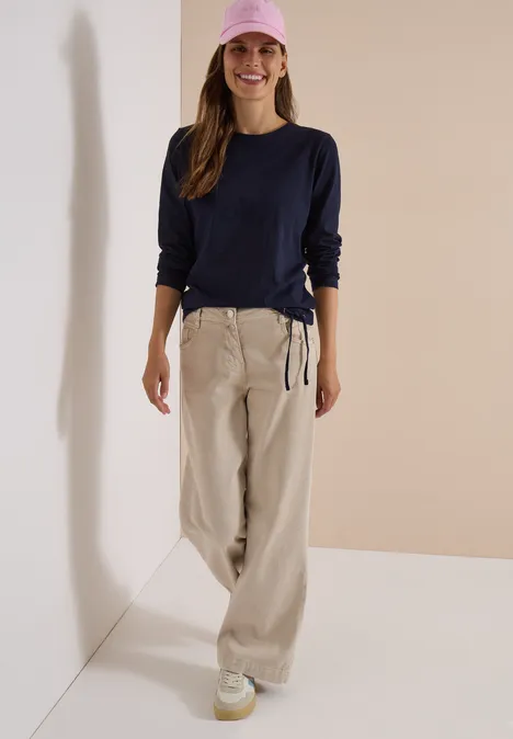 Une femme sourit contre un mur, vêtue d’un sweat bleu marine, d’un pantalon large beige, d’une casquette rose et de baskets.
