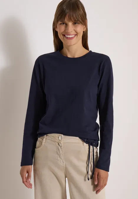 Femme souriante en sweat bleu marine à manches longues et pantalon beige devant un fond clair.