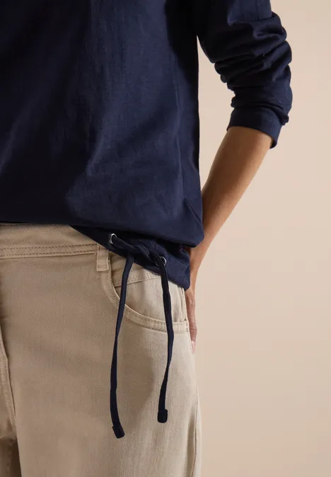 Une personne en pull bleu marine et pantalon crème à cordon, une main dans la poche.