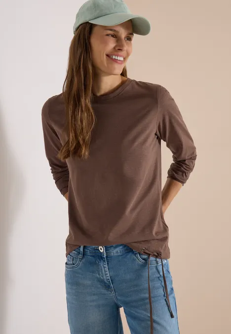 Jeune femme souriante en t-shirt marron, jean et casquette verte, posant de manière décontractée.