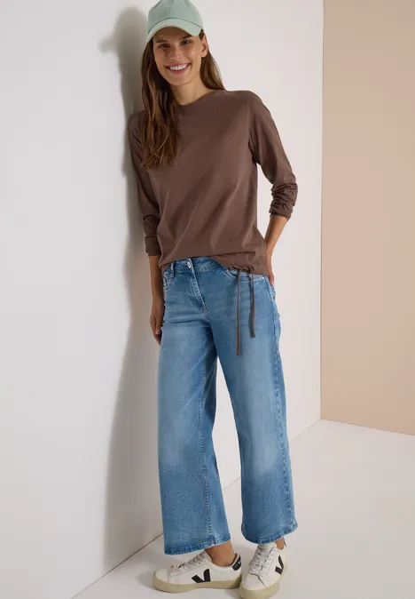 Une femme souriante adossée à un mur, en haut marron, jean large, baskets blanches et casquette verte.