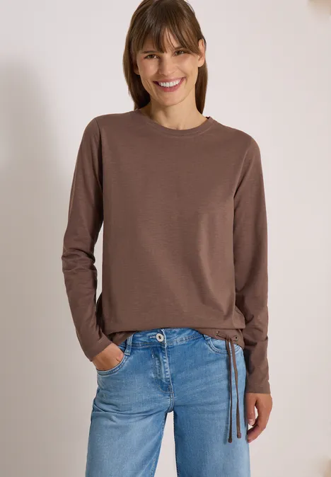 Femme souriante en t-shirt marron à manches longues et jean bleu, posant détendue les mains dans les poches.