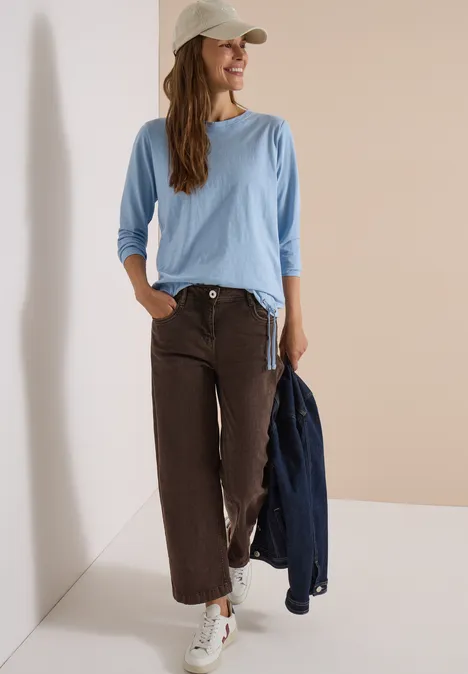 Femme en chemise bleu clair, pantalon large marron et casquette blanche appuyée contre un mur, sourire aux lèvres.