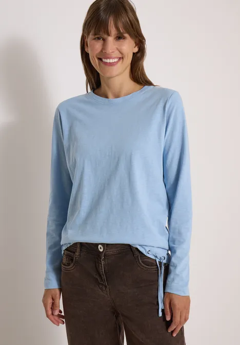 Une femme souriante en haut à manches longues bleu clair et pantalon en velours brun, fond blanc.
