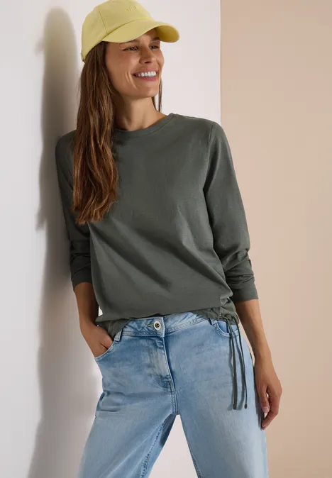 Une femme souriante en tee-shirt vert, jean clair et casquette jaune s’appuie contre un mur.