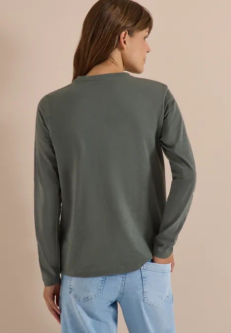 Vue de dos d'une femme portant un haut vert olive à manches longues et un jean bleu clair sur fond neutre.