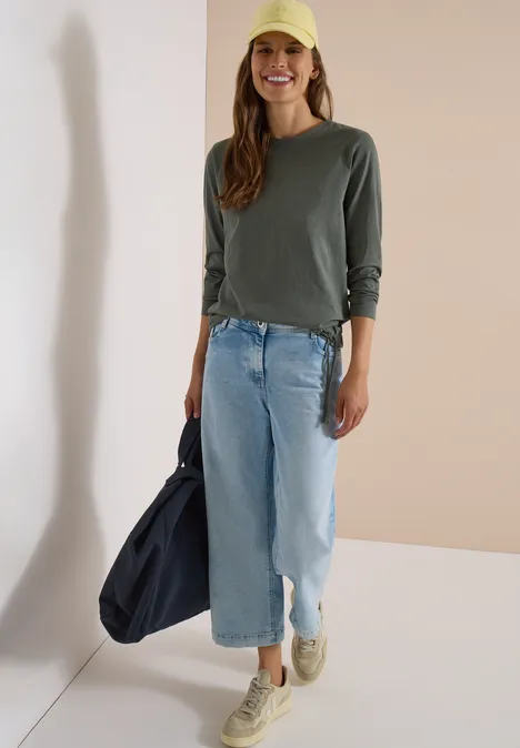Femme en t-shirt vert à manches longues, jean bleu clair, casquette jaune, baskets et sac bleu foncé.