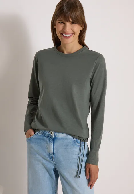 Femme souriante en pull gris à lacets sur le côté et jean bleu clair.