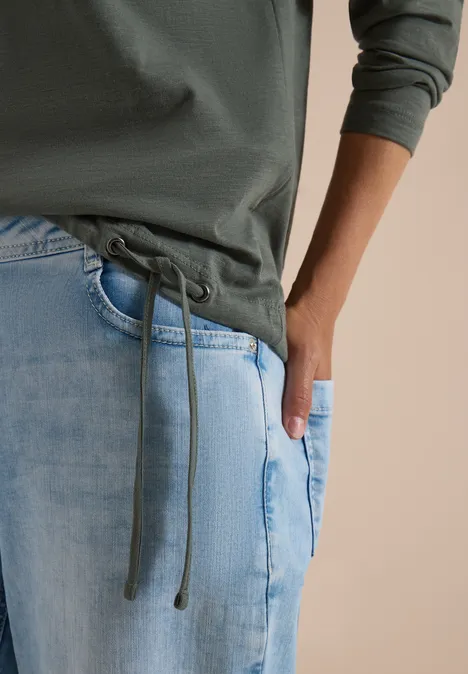 Gros plan d’une personne en sweat à capuche vert à cordons et jean bleu clair, une main dans la poche.