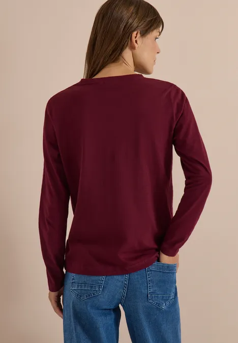 Personne vue de dos portant un t-shirt à manches longues bordeaux et un jean bleu devant un fond uni.