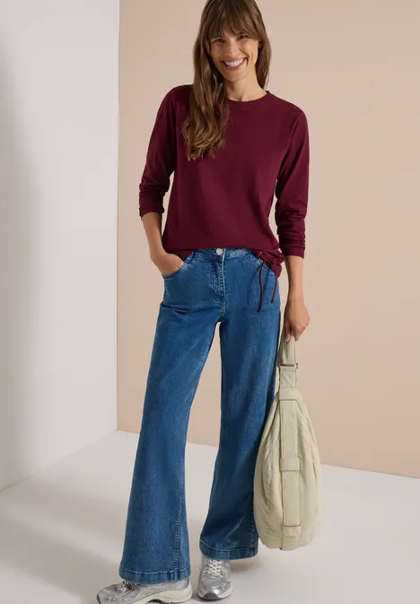 Femme souriante en jean évasé et pull bordeaux, tenant un sac beige.
