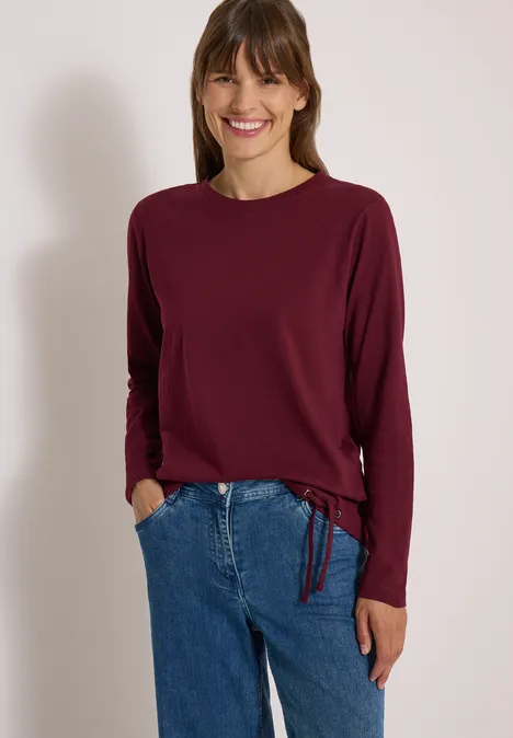 Femme souriante en haut à manches longues bordeaux et jean bleu devant un fond neutre.