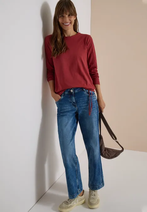 Langarm Shirt mit Rundhals und Tunnelzug russet red Junge Frau lehnt an der Wand, trägt rotes Oberteil, blaue Jeans und eine braune Umhängetasche.