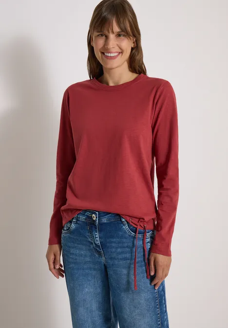 Langarm Shirt mit Rundhals und Tunnelzug russet red Eine lächelnde Frau in rotem Langarmshirt und blauen Jeans vor hellem Hintergrund.