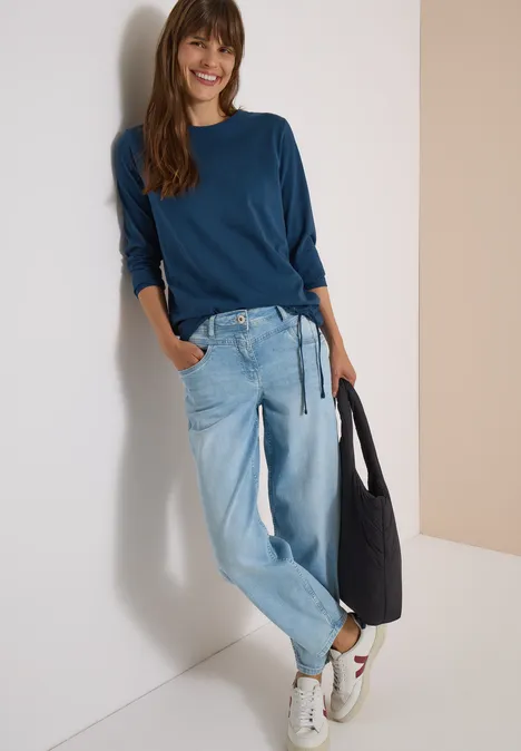 Langarm Shirt mit Rundhals und Tunnelzug moonlight blue Frau lehnt an Wand, trägt blaues Shirt, helle Baggy-Jeans, weiße Sneaker und hält schwarze Tasche.