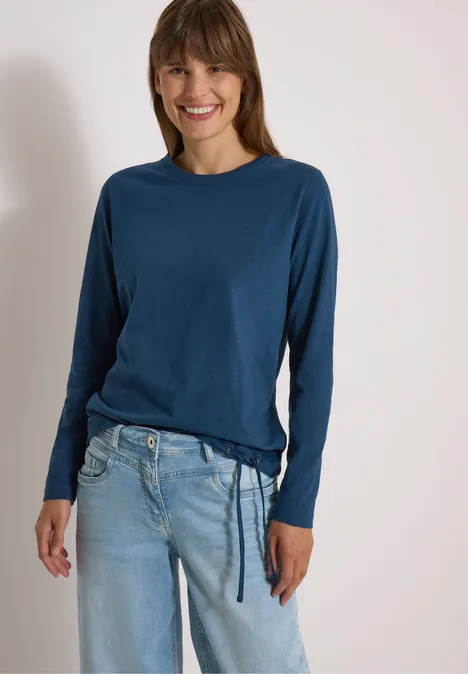 Langarm Shirt mit Rundhals und Tunnelzug moonlight blue Frau in blauem Langarmshirt und hellblauen Jeans lächelt entspannt in die Kamera.