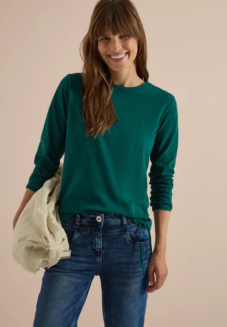 Langarm Shirt mit Rundhals und Tunnelzug dark fir green Eine lächelnde Frau in grünem Pullover und Jeans hält eine beige Jacke vor neutralem Hintergrund.